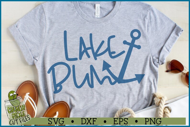 May include: A gray t-shirt with the text "Lake Bum" in blue with an anchor graphic. The shirt is laid out on a wooden surface with sandals and a white tote bag.