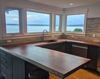 May include: A modern kitchen with a large u-shaped island countertop made of dark wood. The island has a barstool and a sink. The kitchen has large windows with a view of the water.