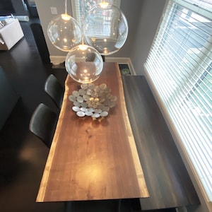 May include: A large wooden dining table with a live edge, black chairs, and a metal bowl filled with silver coins. The table is lit by three clear glass globe pendant lights.