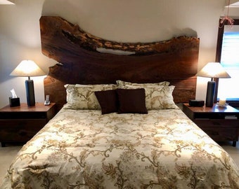May include: A bedroom with a wooden headboard made from a large slab of wood with natural edges. The headboard is a dark brown color and has a unique grain pattern. The bed is made with a white and gold patterned duvet cover and has two bedside tables with lamps.