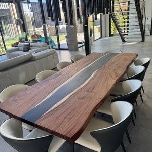 May include: A large wooden dining table with a black epoxy resin inlay. The table has a live edge and is surrounded by white and black chairs.