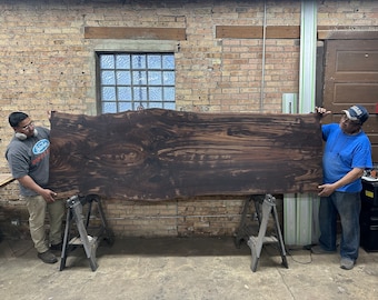 May include: Two people are holding up a large, dark brown wood slab. The slab has a natural edge and a beautiful grain pattern. The slab is resting on two sawhorses.