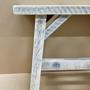 May include: Close-up of a rustic wooden bench with a distressed, weathered finish. The bench features a light gray and white color scheme, with visible wood grain and a sturdy, angled leg design. The bench is designed for indoor or outdoor use.