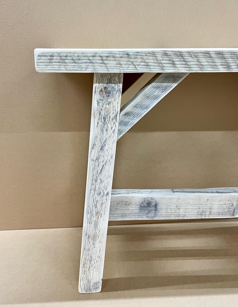 May include: Close-up of a rustic wooden bench with a distressed, weathered finish. The bench features a light gray and white color scheme, with visible wood grain and a sturdy, angled leg design. The bench is designed for indoor or outdoor use.