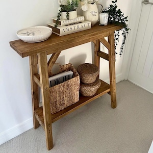 May include: A tall, narrow wooden console table with a dark brown finish, featuring a lower shelf. The top surface holds a white oval dish, two stacked books labelled AESTHETIC and ABSTRACT, a white pitcher, a candle, and a green plant.