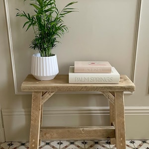 May include: A small, rustic wooden bench with a potted green plant and two books. The white pot has a pleated design. The books are titled "Bohemia" and "Palm Springs". The bench is made of light-colored wood.