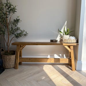 May include: A rustic wooden bench with a natural finish. The bench has a simple design with a slatted lower shelf. A small decorative item sits on the bench. A potted plant and a woven basket with a plant are on either side of the bench.