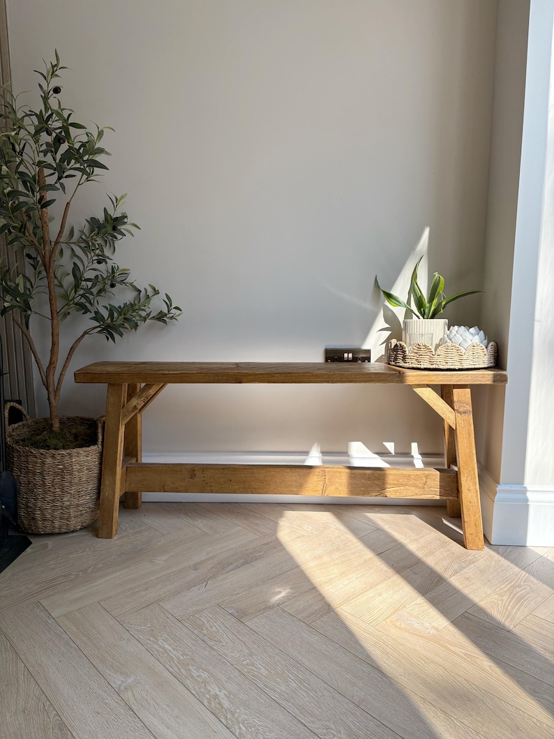 May include: A rustic wooden bench with a natural finish. The bench has a simple design with a slatted lower shelf. A small decorative item sits on the bench. A potted plant and a woven basket with a plant are on either side of the bench.