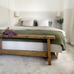 May include: A bedroom scene featuring a bed with a light gray headboard, olive green bedding, and cream-colored pillows. A wooden bench sits at the foot of the bed, and a vase of greenery is on the side table. The walls are painted in neutral tones.