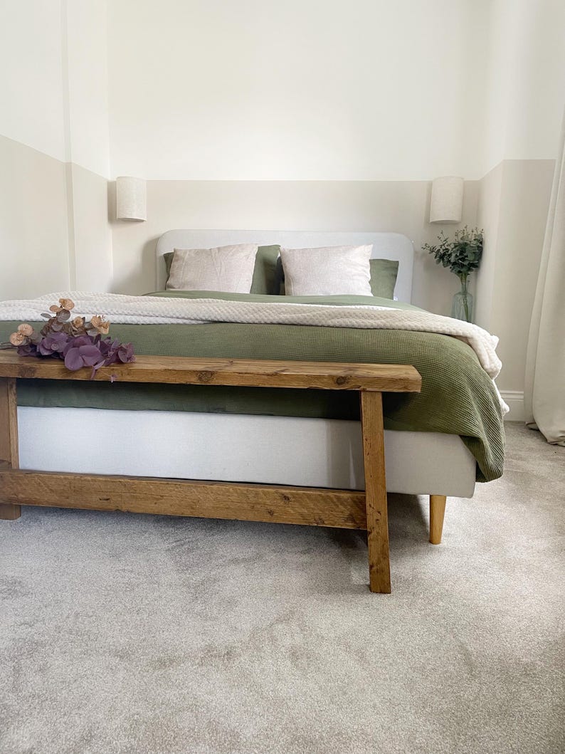 May include: A bedroom scene featuring a bed with a light gray headboard, olive green bedding, and cream-colored pillows. A wooden bench sits at the foot of the bed, and a vase of greenery is on the side table. The walls are painted in neutral tones.