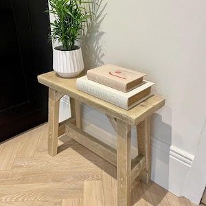 May include: A small, light-colored wooden bench with a rectangular top and four sturdy legs. Two books, "Bohemia" and "Palm Springs," are stacked on the bench. A potted plant with green leaves sits on the bench. The bench is made of natural wood.