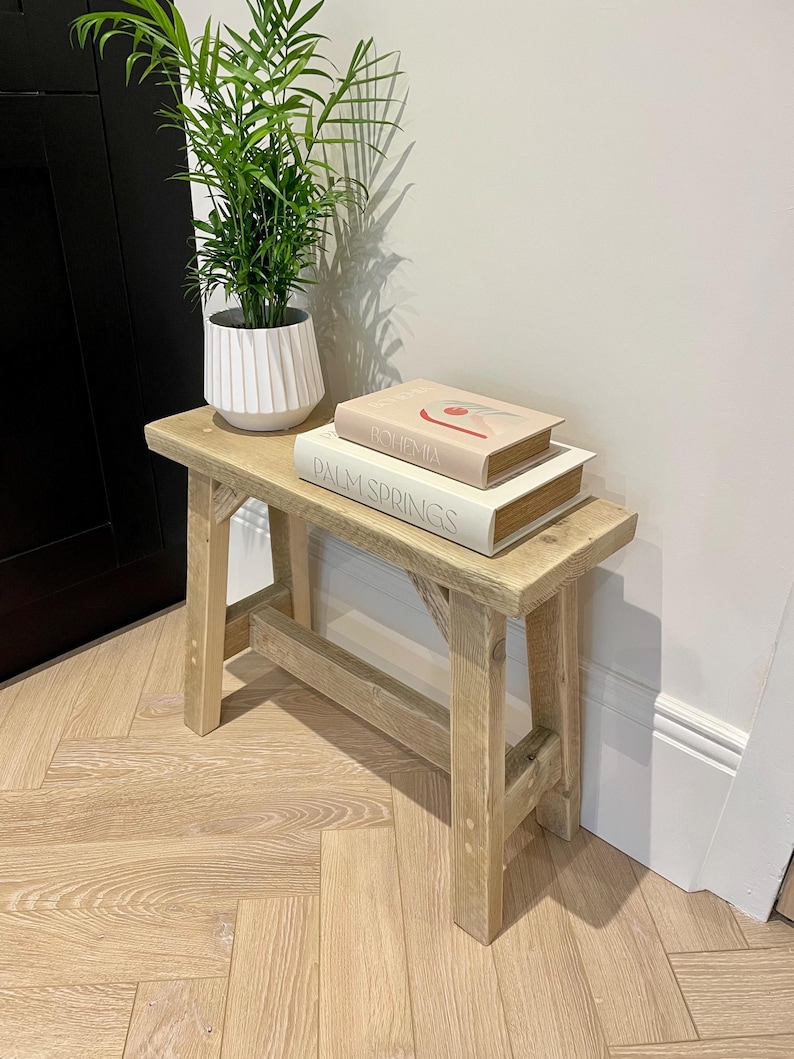 May include: A small, light-colored wooden bench with a rectangular top and four sturdy legs. Two books, "Bohemia" and "Palm Springs," are stacked on the bench. A potted plant with green leaves sits on the bench. The bench is made of natural wood.
