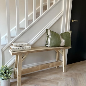 May include: A rustic wooden bench with a green and white striped pillow and two books that read "Aesthetic Abstract." A small potted plant sits to the left of the bench. The background includes a staircase and a black door.