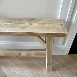 May include: A rustic wooden bench with a natural, weathered finish. The bench features a rectangular seat supported by angled legs, showcasing visible wood grain and knots. The light brown color and simple design suggest a farmhouse or cottage style.