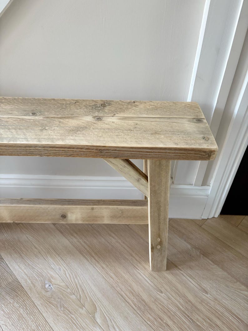 May include: A rustic wooden bench with a natural, weathered finish. The bench features a rectangular seat supported by angled legs, showcasing visible wood grain and knots. The light brown color and simple design suggest a farmhouse or cottage style.