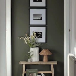May include: A hallway scene featuring three framed monochrome photographs on a dark green wall. A wooden bench below displays a white vase with flowers, a small lamp, a potted plant, and a woven basket with a book and flowers.