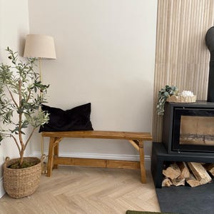 May include: A rustic wooden bench with a black velvet pillow sits in a cozy living space. A floor lamp with a beige shade and a potted olive tree in a woven basket add to the room's decor. A fireplace with stacked firewood is visible.