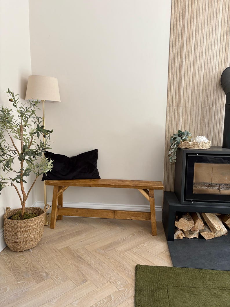 May include: A rustic wooden bench with a black velvet pillow sits in a cozy living space. A floor lamp with a beige shade and a potted olive tree in a woven basket add to the room's decor. A fireplace with stacked firewood is visible.
