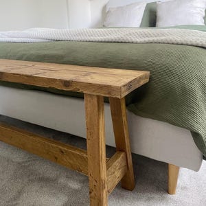 May include: A rustic wooden bench with a natural finish. The bench has a rectangular seat and angled legs, showcasing the wood grain. The bench is placed in front of a bed with a green blanket and white pillows.