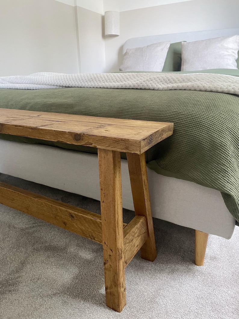 May include: A rustic wooden bench with a natural finish. The bench has a rectangular seat and angled legs, showcasing the wood grain. The bench is placed in front of a bed with a green blanket and white pillows.