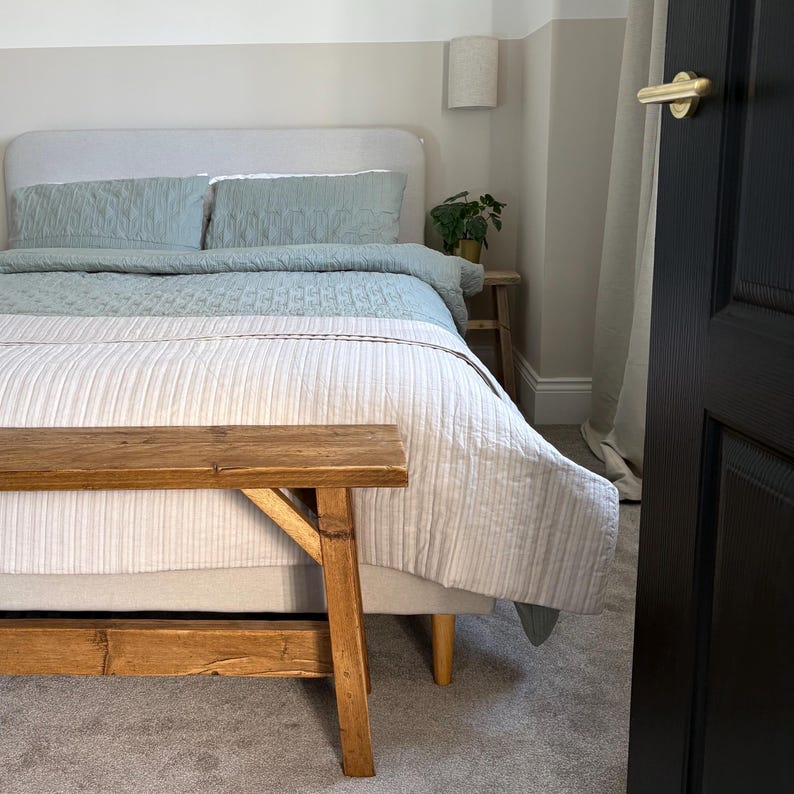 May include: A bedroom scene featuring a bed with a light blue quilt and pillows, and a white and beige striped comforter. A wooden bench sits at the foot of the bed. The room has a neutral color palette.