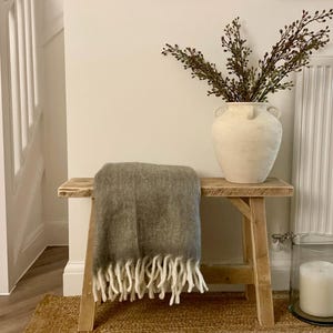 May include: A rustic wooden bench with a gray throw blanket and white fringe. A large cream-colored vase holds leafy branches. The bench sits on a woven jute rug, creating a cozy, neutral-toned interior scene.