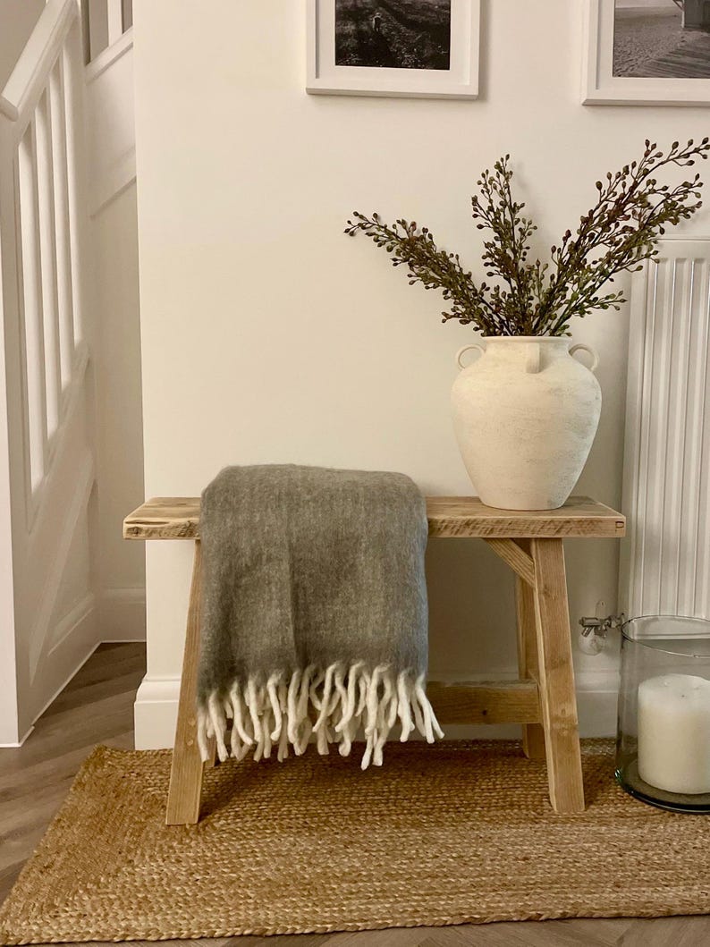 May include: A rustic wooden bench with a gray throw blanket and white fringe. A large cream-colored vase holds leafy branches. The bench sits on a woven jute rug, creating a cozy, neutral-toned interior scene.