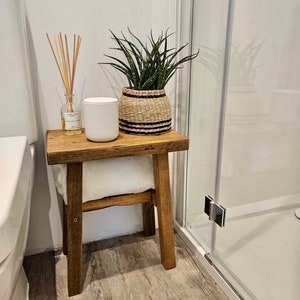 May include: A small, rustic wooden stool with a white towel underneath. On top, a potted snake plant in a woven basket with black stripes, a white candle, and a glass diffuser bottle with reeds. The stool is in a bathroom setting.
