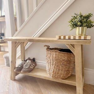 May include: A light-toned wooden console table with a lower shelf, featuring a woven seagrass basket, a pair of brown and white trainers, a gold-coloured planter with a green plant, and a rectangular tray with a white and tan striped pattern.