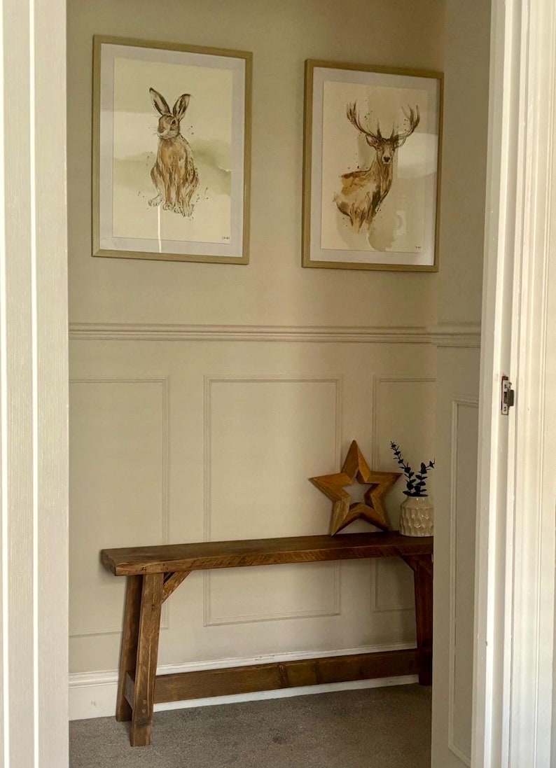 May include: A hallway with framed watercolor art of a hare and a deer above a wooden bench. A wooden star and a small vase with greenery are on the bench. The walls are painted a light beige.