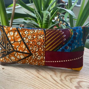 May include: A rectangular, patchwork fabric pouch with a blue zip. The pouch features vibrant patterns in orange, brown, burgundy, and blue. The pouch is resting on a wooden surface, with green plants in the background.