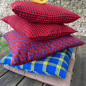 May include: Four cushions stacked on top of each other. The top two are red and black gingham. The third is burgundy with a blue bow tie design. The bottom cushion is blue tartan with yellow stripes.