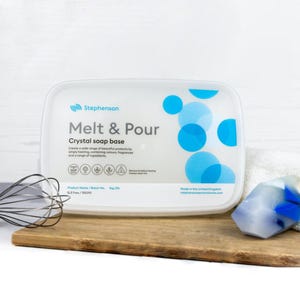 May include: A white container of Stephenson Melt & Pour Crystal soap base, with blue circular designs and text. A whisk and a piece of soap are on a wooden board. Made in the United Kingdom.