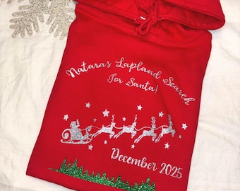 Può includere: Una felpa con cappuccio rossa con la scritta "Natara's Lapland Search for Santa!" in glitter argento. Il design include una slitta glitterata argento con renne, stelle e alberi glitterati verdi. È presente anche la data "Dicembre 2025".