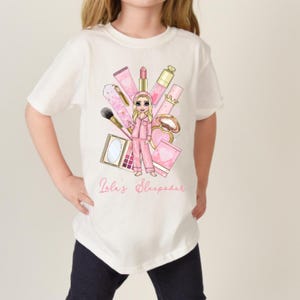 Op de afbeelding: Een wit t-shirt met een grafische illustratie van een blond personage in een roze pyjama, omringd door diverse make-up artikelen zoals kwasten, lippenstift en paletten. De tekst "Lola's Sleepover" is in roze letters onder de afbeelding gedrukt.
