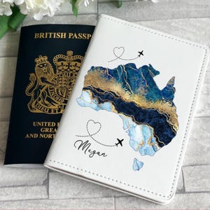 May include: A white passport holder featuring a blue and gold map design, airplane and heart graphics, and the name "Megan". A dark blue British passport is next to it. A travel accessory.