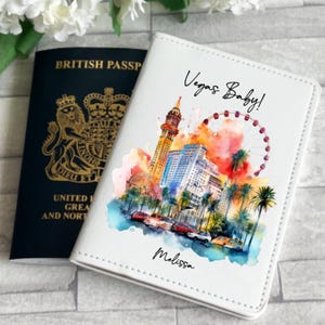 Puede incluir: Una funda blanca para pasaporte con una colorida ilustración de acuarela de los lugares emblemáticos de Las Vegas, incluyendo la réplica de la Torre Eiffel y una noria. La funda está personalizada con el nombre "Melissa" y la frase "Vegas Baby!" Un pasaporte británico está al lado.