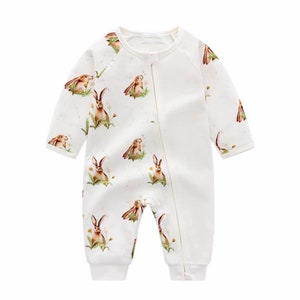 May include: White baby romper with a full-length zipper and long sleeves. The romper features a repeating pattern of brown and tan rabbits in a field of green grass and yellow flowers. The fabric has a speckled, watercolor-like effect.