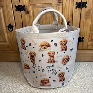May include: A beige felt toy basket with a rounded shape and a handle. The basket is decorated with images of brown puppies, black hearts, and bone shapes. The text "Rolo's Toys" is printed on the front.