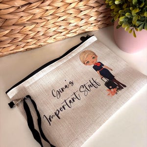 May include: A light-colored canvas zippered pouch with a black zipper and wrist strap. The pouch features an illustration of a woman in a uniform with a suitcase and the text "Gina's Important Stuff" in black script.