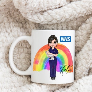 Puede incluir: Taza de cerámica blanca con una ilustración de dibujos animados de una enfermera sosteniendo a un bebé frente a un arcoíris. La taza tiene el texto "NHS" y el nombre "Katie". La enfermera lleva un uniforme azul marino.