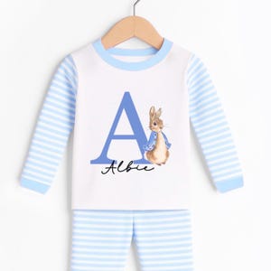 May include: A white and light blue striped pajama set. The top features a large blue letter "A" and a watercolor illustration of a rabbit wearing a blue jacket. The name "Albie" is written in cursive below the letter.
