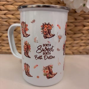 Op de afbeelding: Witte emaille mok met een zilveren rand en handvat. De mok heeft een herhalend patroon van bruine cowboylaarzen gevuld met oranje en roze bloemen, met de tekst "Sorta Sweet Sorta Beth Dutton."
