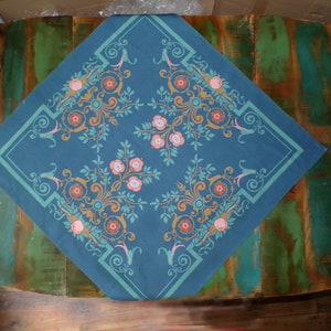 Könnte beinhalten: Ein quadratisches, teal-farbenes Tischtuch mit floralem Design. Das Design zeigt rosa Rosen, orangefarbene und tealfarbene Akzente und einen tealfarbenen Rand. Das Tischtuch liegt auf einem alten, mehrfarbigen Holztisch.