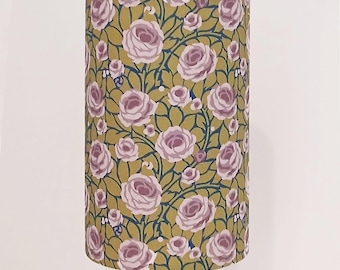 May include: Cylindrical lampshade featuring a repeating pattern of lavender roses and green leaves on a mustard yellow background. The floral design is set against a dark blue vine-like pattern. The lampshade is mounted on a copper-colored stand.