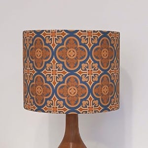 May include: A decorative lampshade with a repeating floral pattern in shades of brown, orange, and blue. The lampshade is cylindrical and sits atop a wooden lamp base. The design is reminiscent of vintage or retro styles.