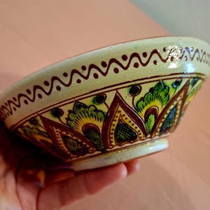 Puede incluir: Un cuenco de cerámica con base de color crema y un patrón de zigzag marrón pintado a mano en el borde. El exterior está decorado con un motivo repetitivo de hojas puntiagudas verdes y amarillas delineadas con esmalte marrón oscuro.