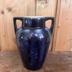 Op de afbeelding: Een donkerblauwe keramische vaas met een glanzende afwerking en een gevlekt wit patroon. De vaas heeft twee zwarte handvatten aan weerszijden, een brede hals en een goudkleurige rand. Het lichaam van de vaas heeft een ronde vorm die naar onderen toe afloopt.