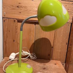 Peut inclure: Une lampe de bureau vert citron avec un col flexible argenté et une base ronde. L'abat-jour a des découpes blanches en forme de nuage. La lampe est branchée sur une prise électrique blanche. La lampe est sur une surface en bois.