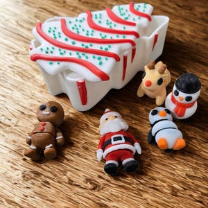 Op de afbeelding: Een witte kerstboomvormige container met rode en groene accenten, omringd door miniatuur vakantiefiguren. De figuren omvatten een peperkoekmannetje, de kerstman, een rendier, een sneeuwpop en een pinguïn. Op een houten oppervlak.